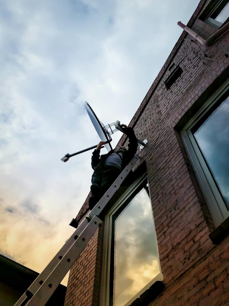 A technician balances on a ladder while installing a satellite dish on an exterior brick wall at dusk.